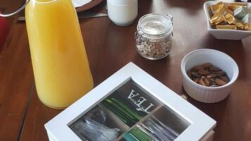A breakfast table with a bottle of orange juice, a glass, a basket of bread, a bowl of almonds, and a box of tea.
