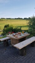 An outdoor picnic setup with a wooden table and benches, surrounded by greenery and a view of a field.