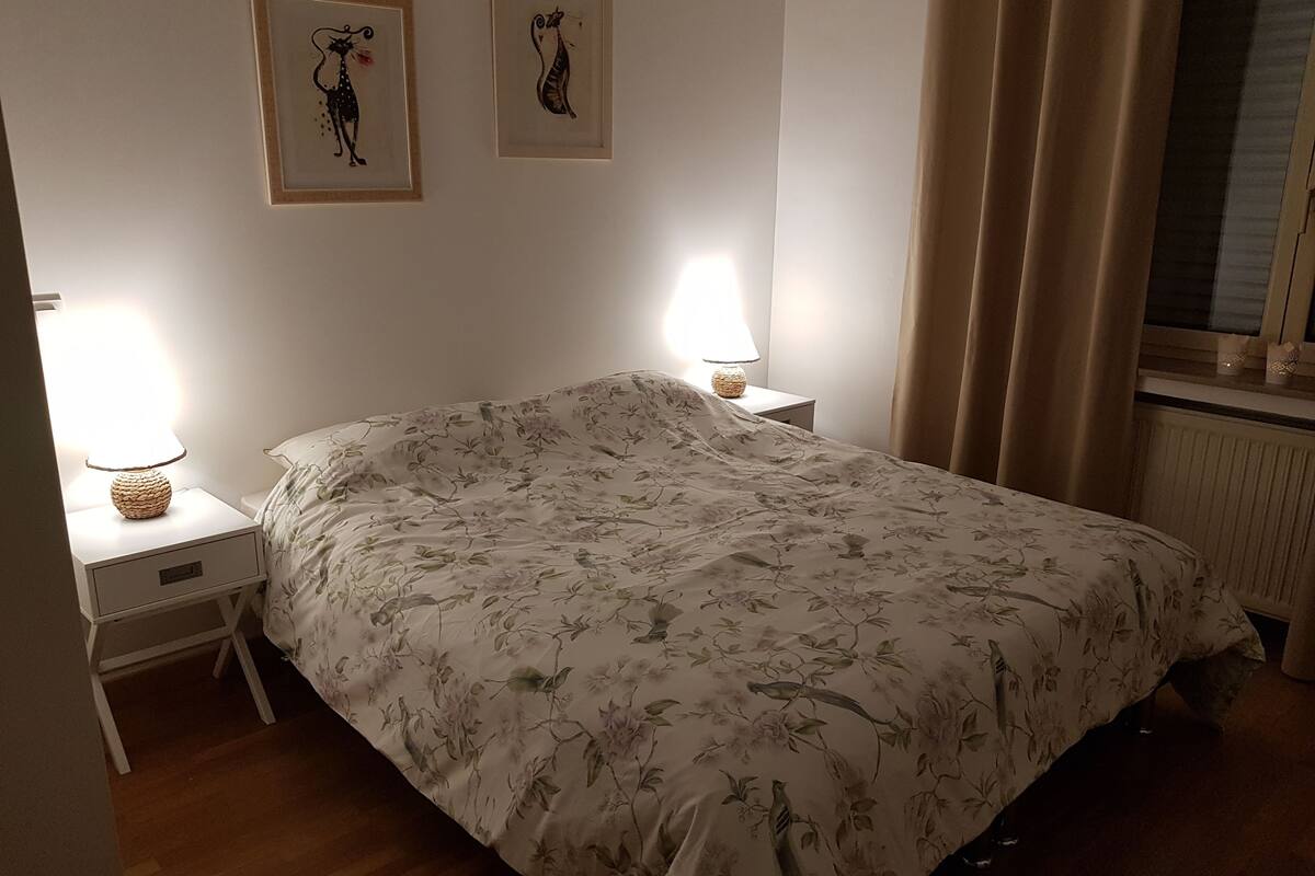 A bedroom with a floral bedspread, two wall-mounted picture frames, bedside lamps, a window with curtains, and a radiator.