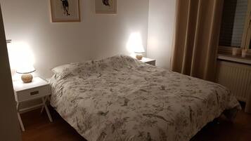 Une chambre à coucher avec une couvre-lit à motifs floraux, deux cadres photos fixés au mur, des lampes de chevet, une fenêtre avec des rideaux et un radiateur.