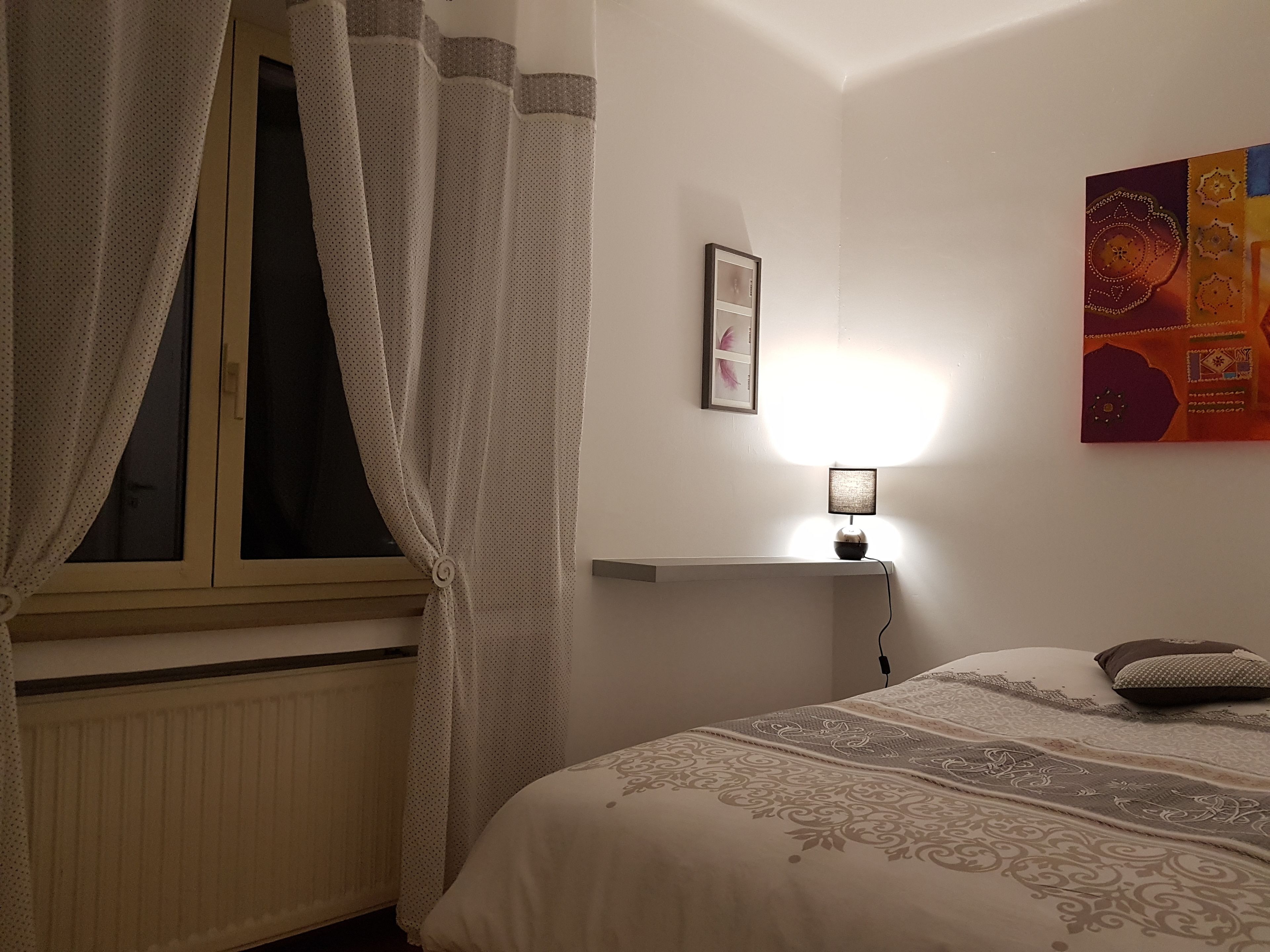 Une chambre à coucher avec un lit, une fenêtre avec des rideaux, une lampe de chevet, une étagère fixée au mur et une affiche.