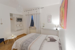 A bedroom with a bed, a TV on a shelf, a small table, and a window with curtains.
