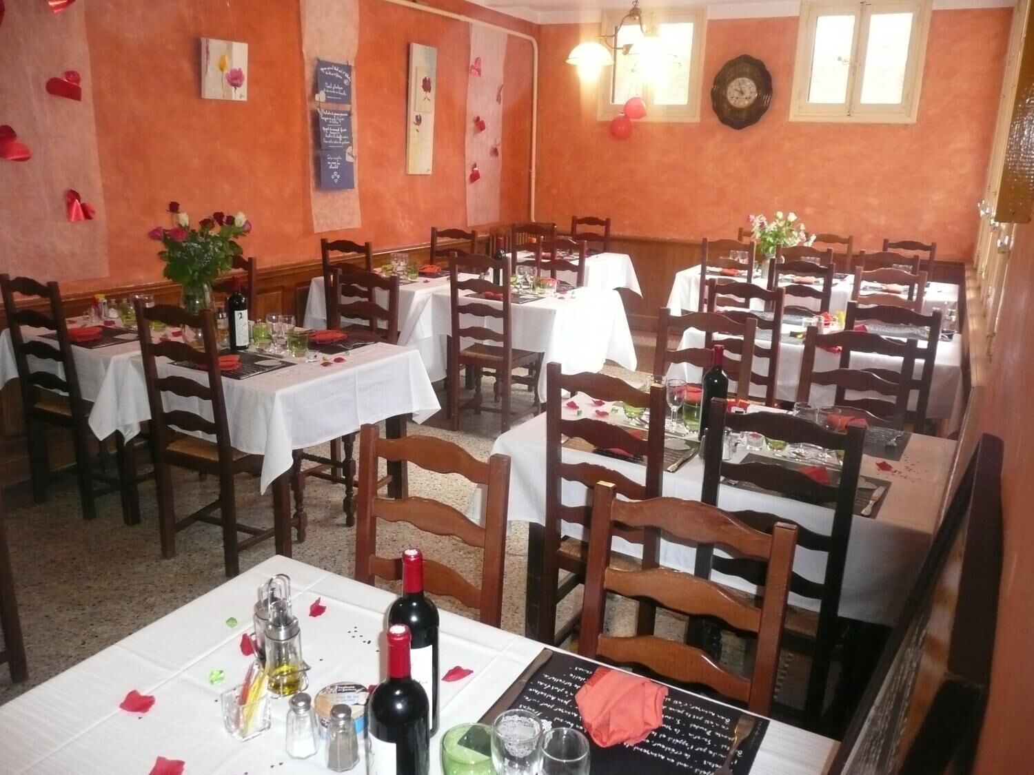 A restaurant with multiple tables, each set with white tablecloths, wooden chairs, and red rose petals.