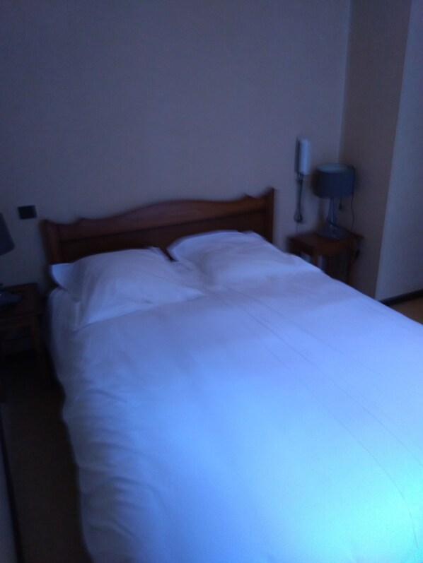 Una habitación de hotel estándar con una cama de madera, una mesita de noche con lámpara y un teléfono fijo en la pared.