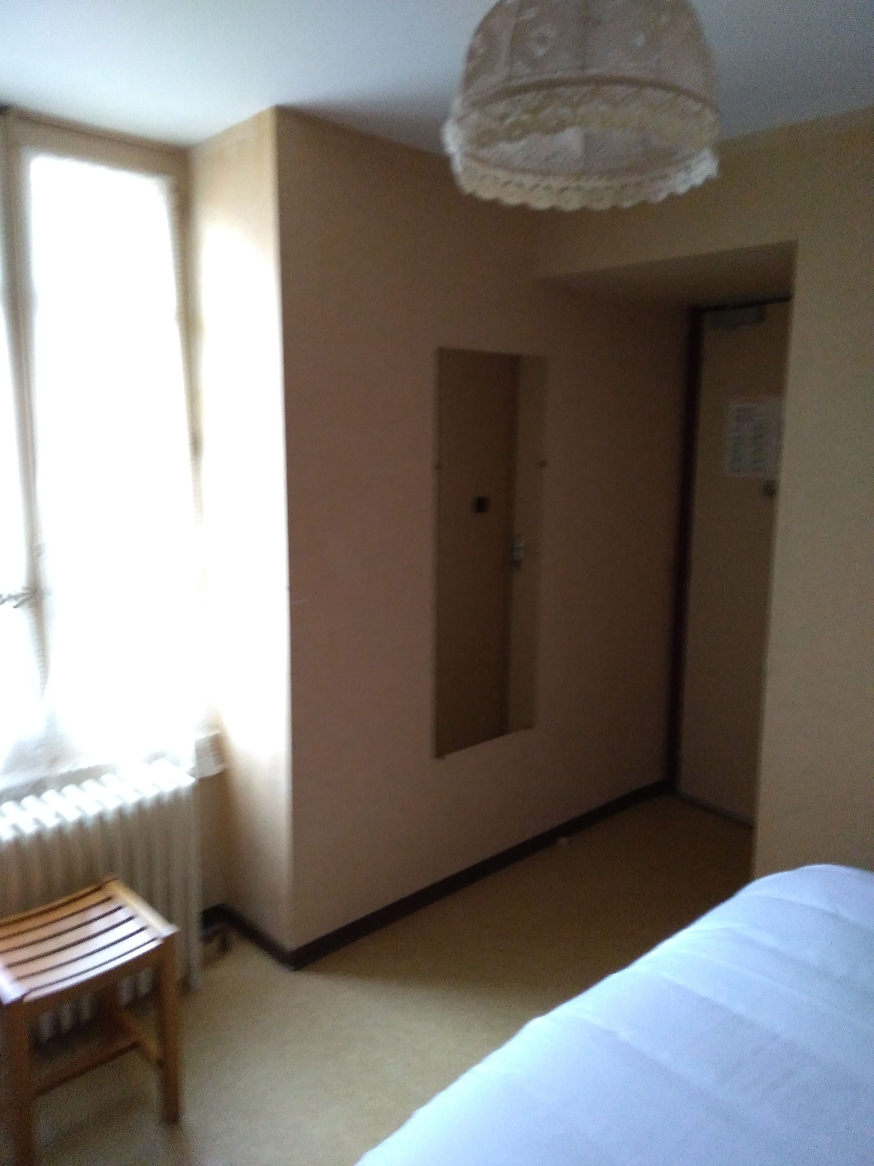 A hotel room with a bed, a wooden chair, a radiator, a door, and a light fixture.
