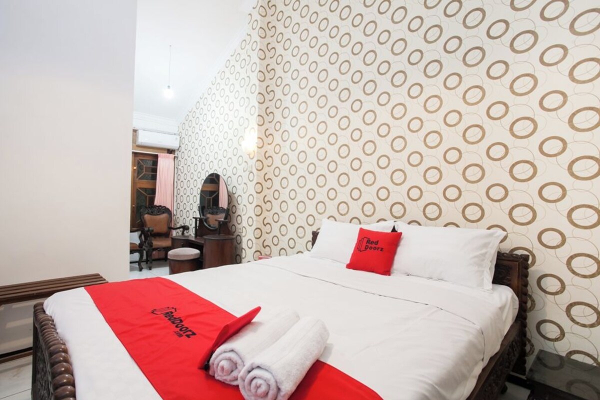 Une chambre d’hôtel avec un lit, une couvre-lit rouge et blanche, un oreiller rouge et un mur orné d’un motif circulaire.