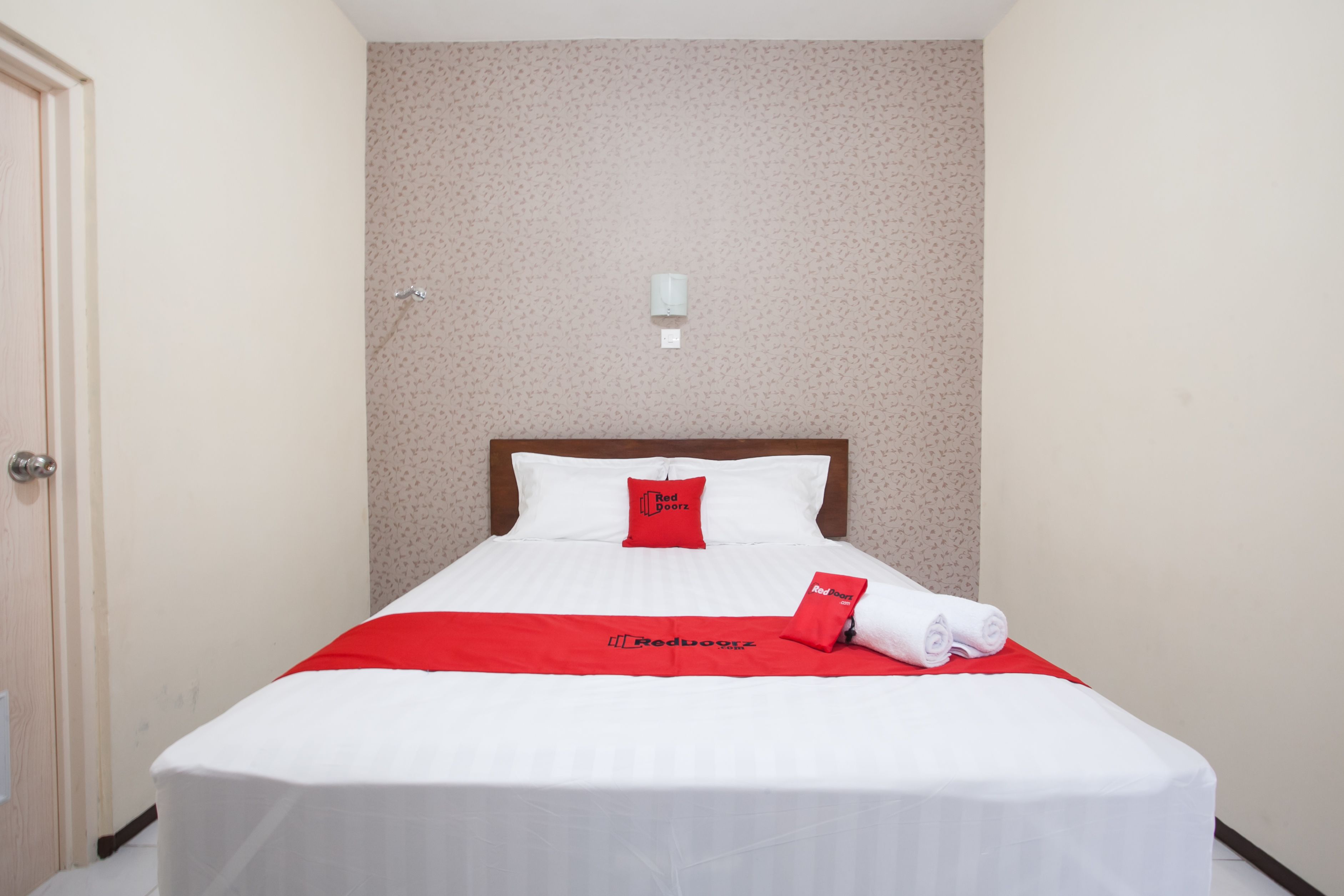 A neatly made bed with white bedding and red accents, a wall-mounted light fixture, and a door to the left.
