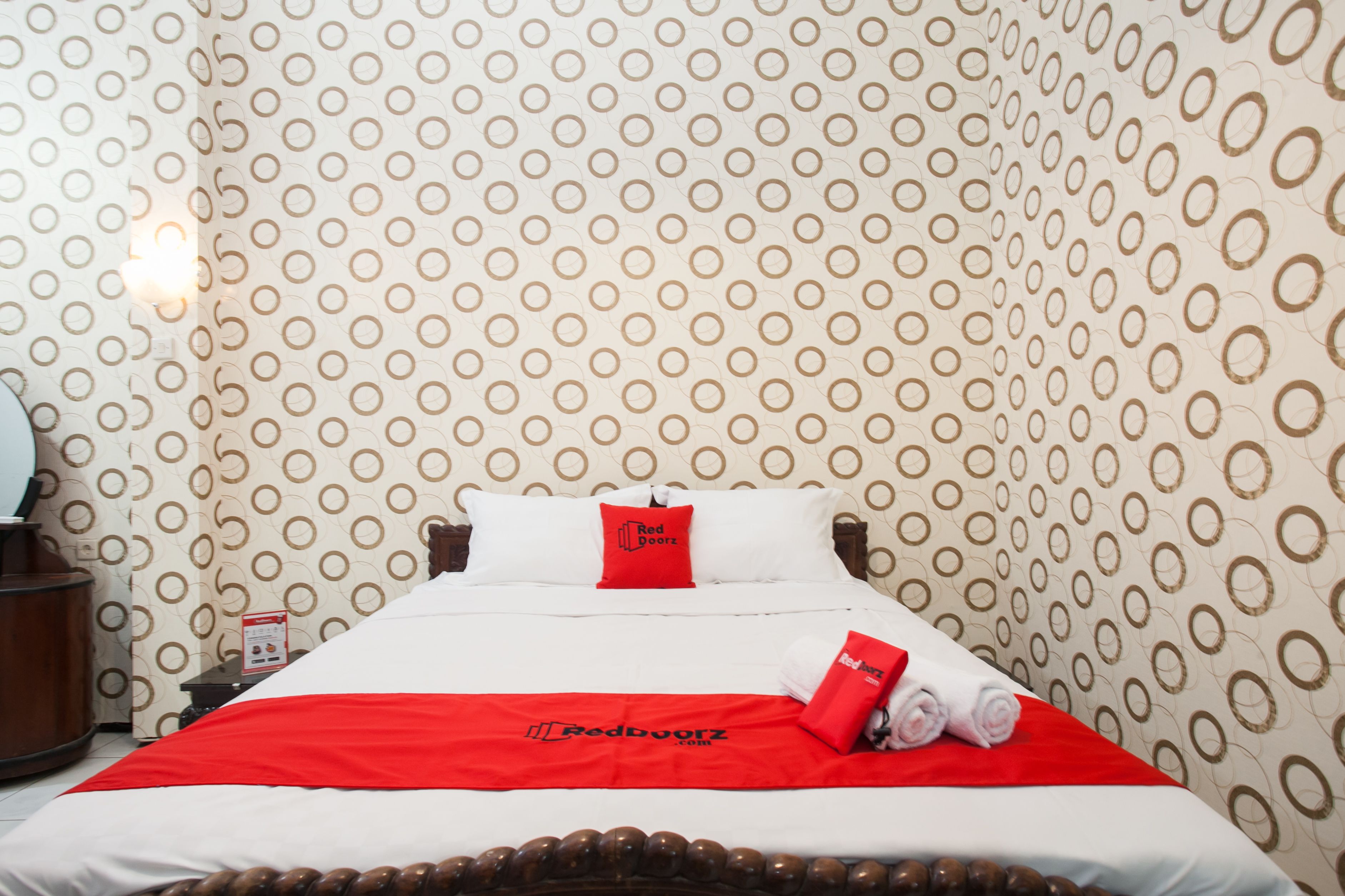 Ein Hotelzimmer mit ornamental gestalteter Tapete, einem ordentlich bezogenen Bett mit rotem und weißem Überwurf und einem roten Kopfkissen mit dem Hotel-Logo.