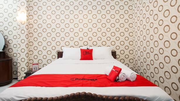 Ein Hotelzimmer mit ornamental gestalteter Tapete, einem ordentlich bezogenen Bett mit rotem und weißem Überwurf und einem roten Kopfkissen mit dem Hotel-Logo.