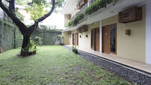 A courtyard with a tree, potted plants, a pathway, and a building with balconies and wooden doors. - RedDoorz Plus near Cilandak Town Square 2 (Jakarta)