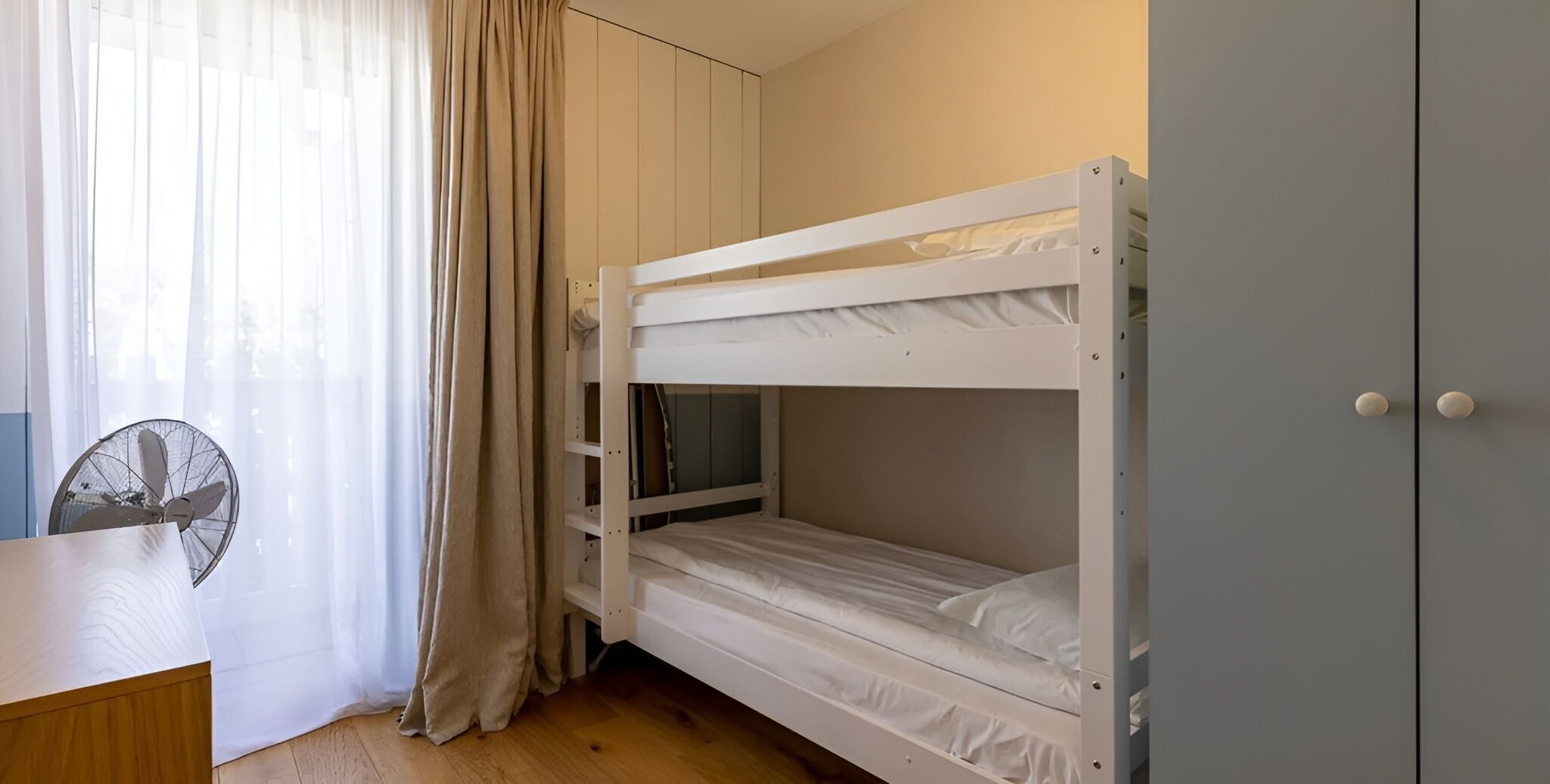 A bunk bed room with a desk, a fan, and a wardrobe.
