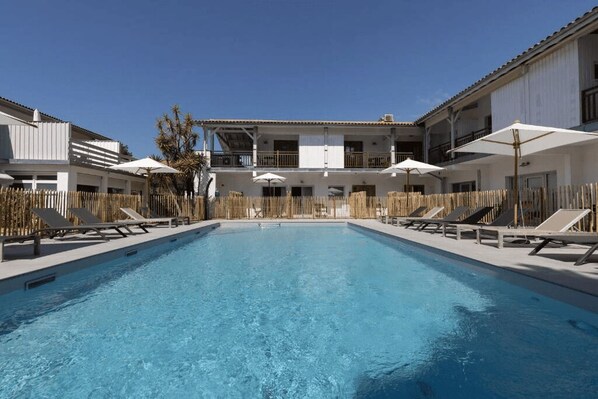 A swimming pool with lounge chairs and umbrellas, surrounded by a wooden fence and buildings with balconies.
