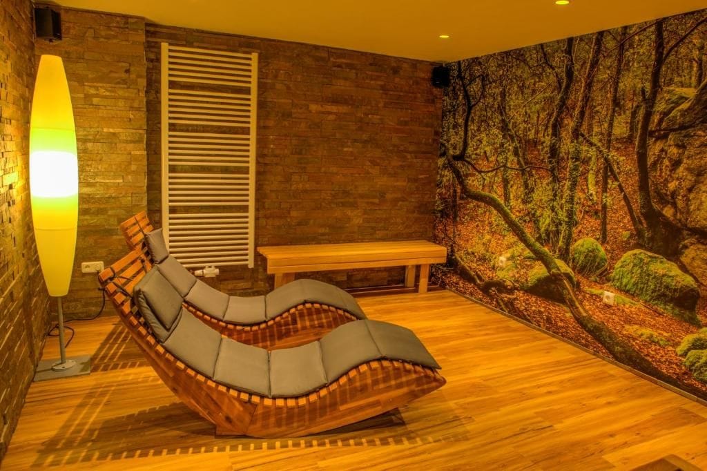 A room with a curved wooden lounge chair, a bench, a wall-mounted heater, and a forest-themed mural.