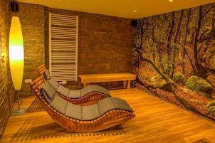 Una habitación con una butaca de madera curvada, un banco, un calentador fijado a la pared y un mural con motivos de bosque.