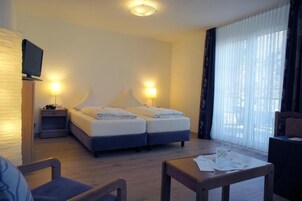 Quarto de hotel com cama, abajures, televisão em um móvel, escrivaninha com papéis e janela com cortinas.