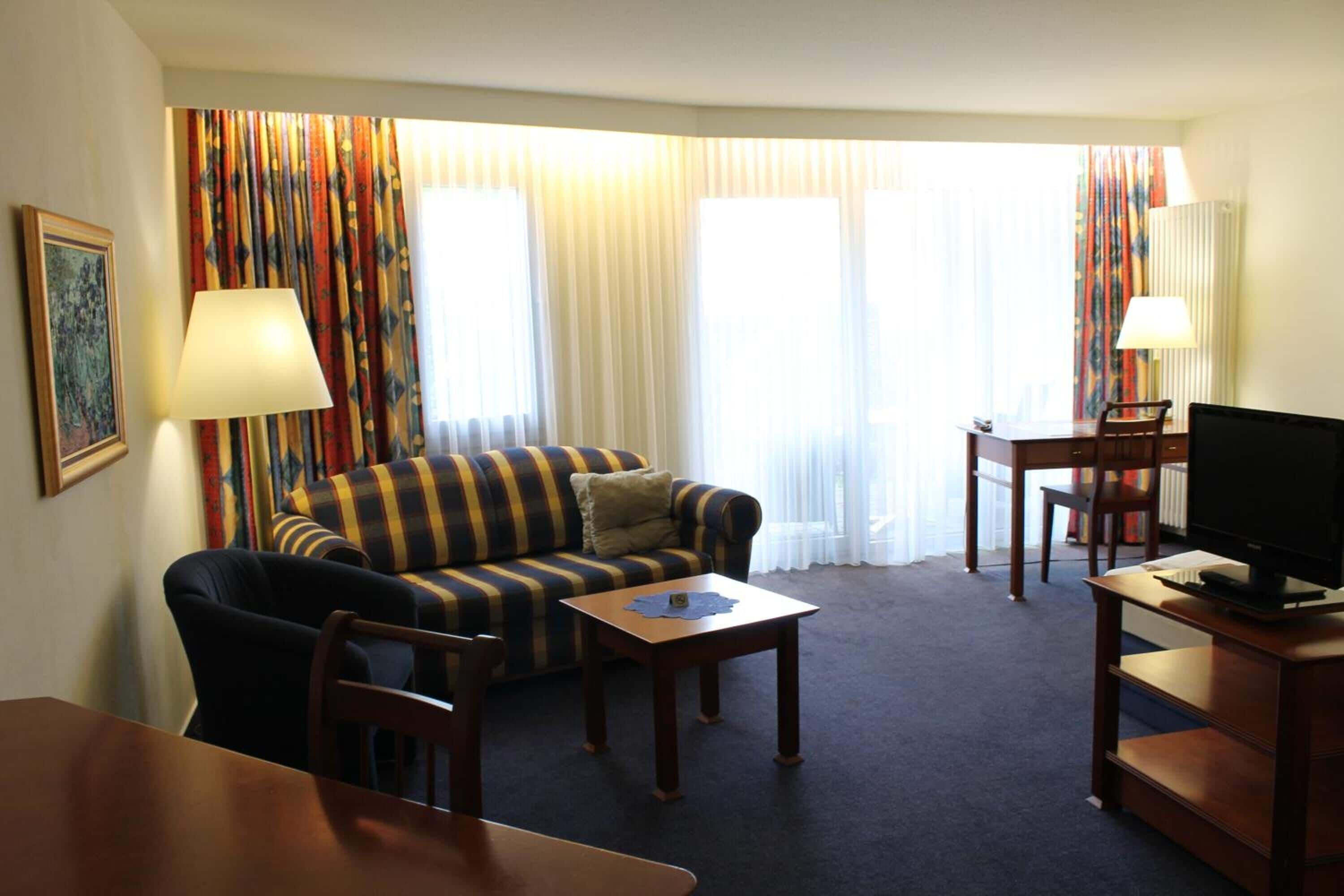 A hotel room with a striped sofa, a small table, a TV on a wooden stand, and a lamp.