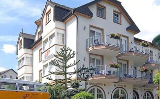 Um hotel de vários andares com varandas, um letreiro visível para 'Haus Apollo Lounge' e um ônibus estacionado.