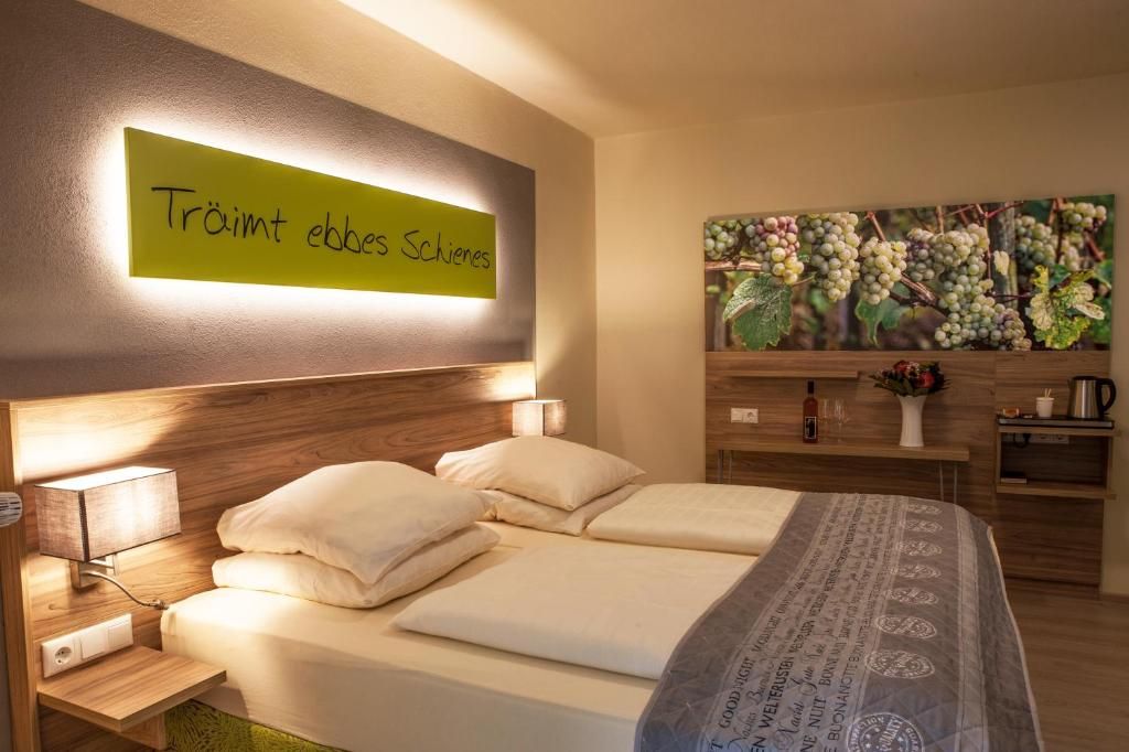 Una habitación de hotel con una cama, mesitas de noche, una lámpara de noche, un cartel en la pared y una pintura de uvas.