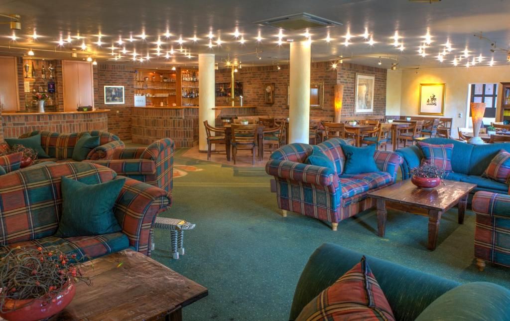 A cozy lounge area with plaid sofas and wooden tables, a bar with shelves of bottles, and a brick wall backdrop.