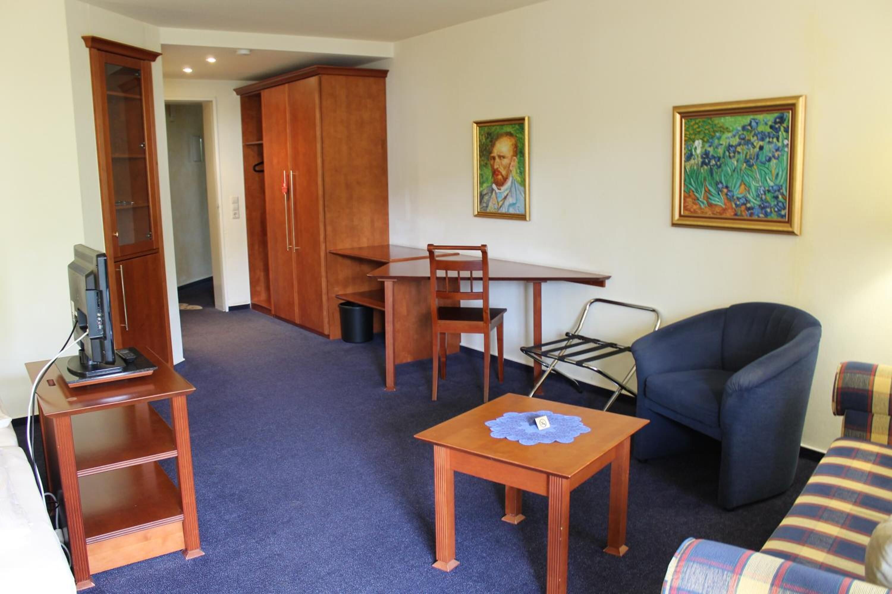 Una sala de estar con un televisor sobre un soporte de madera, una mesa de comedor con sillas, un sillón azul y un sofá con funda a cuadros.