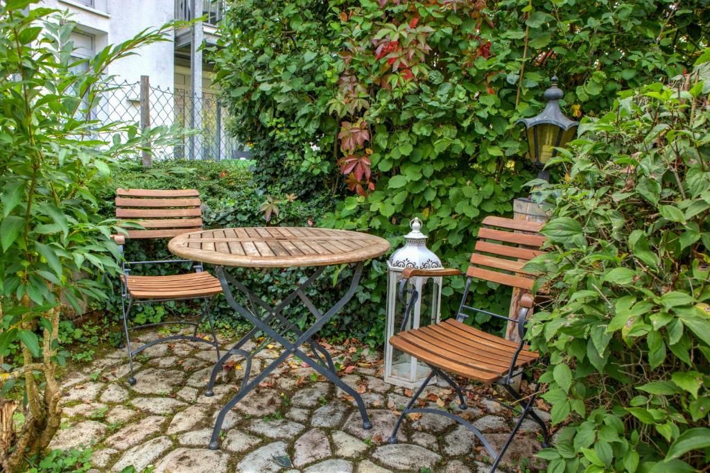 An outdoor seating area with a round table and four wooden chairs, surrounded by lush greenery and a cobblestone floor.