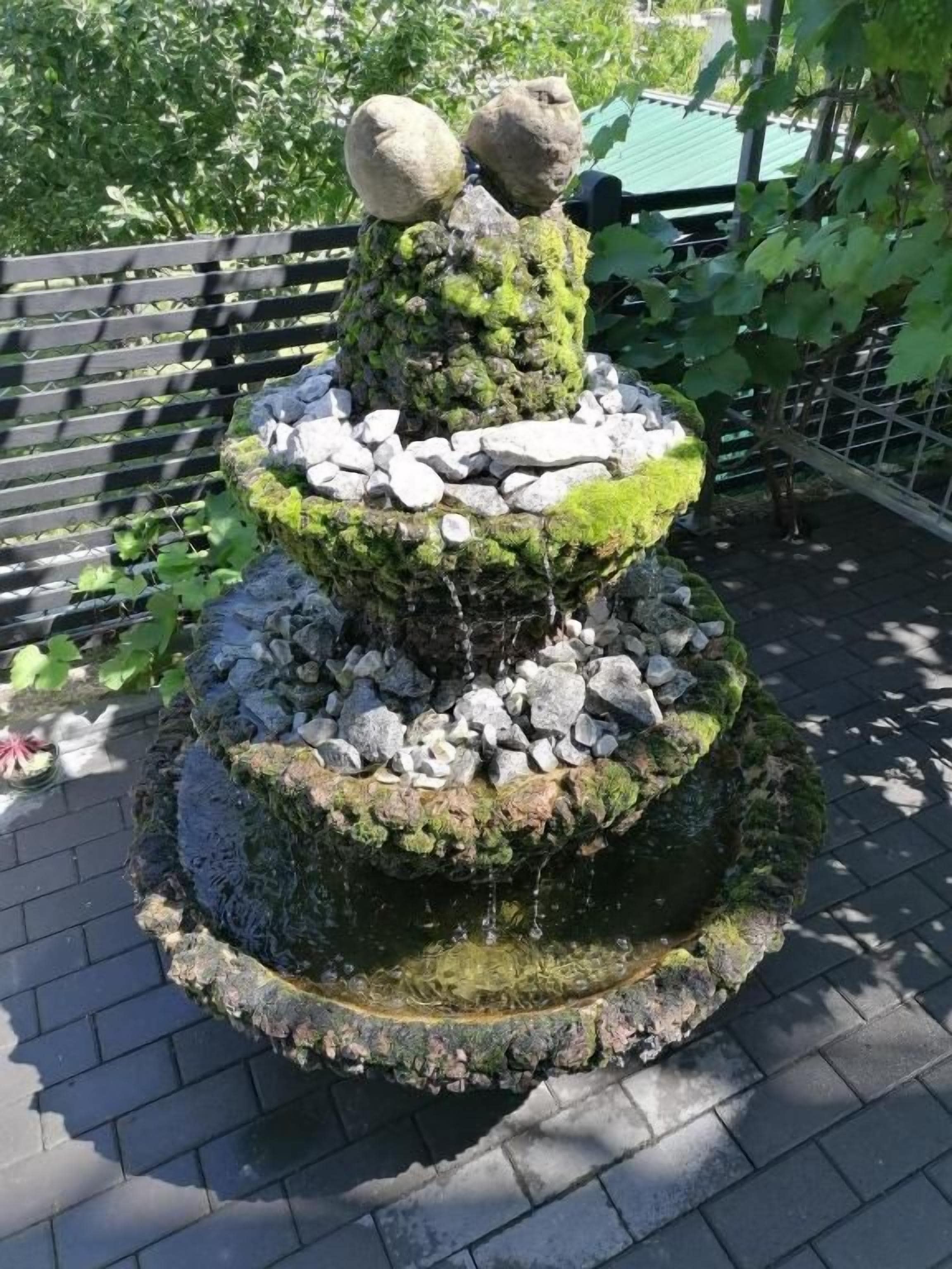 Una fuente de piedra escalonada cubierta de musgo y rocas, rodeada por una cerca y follaje.