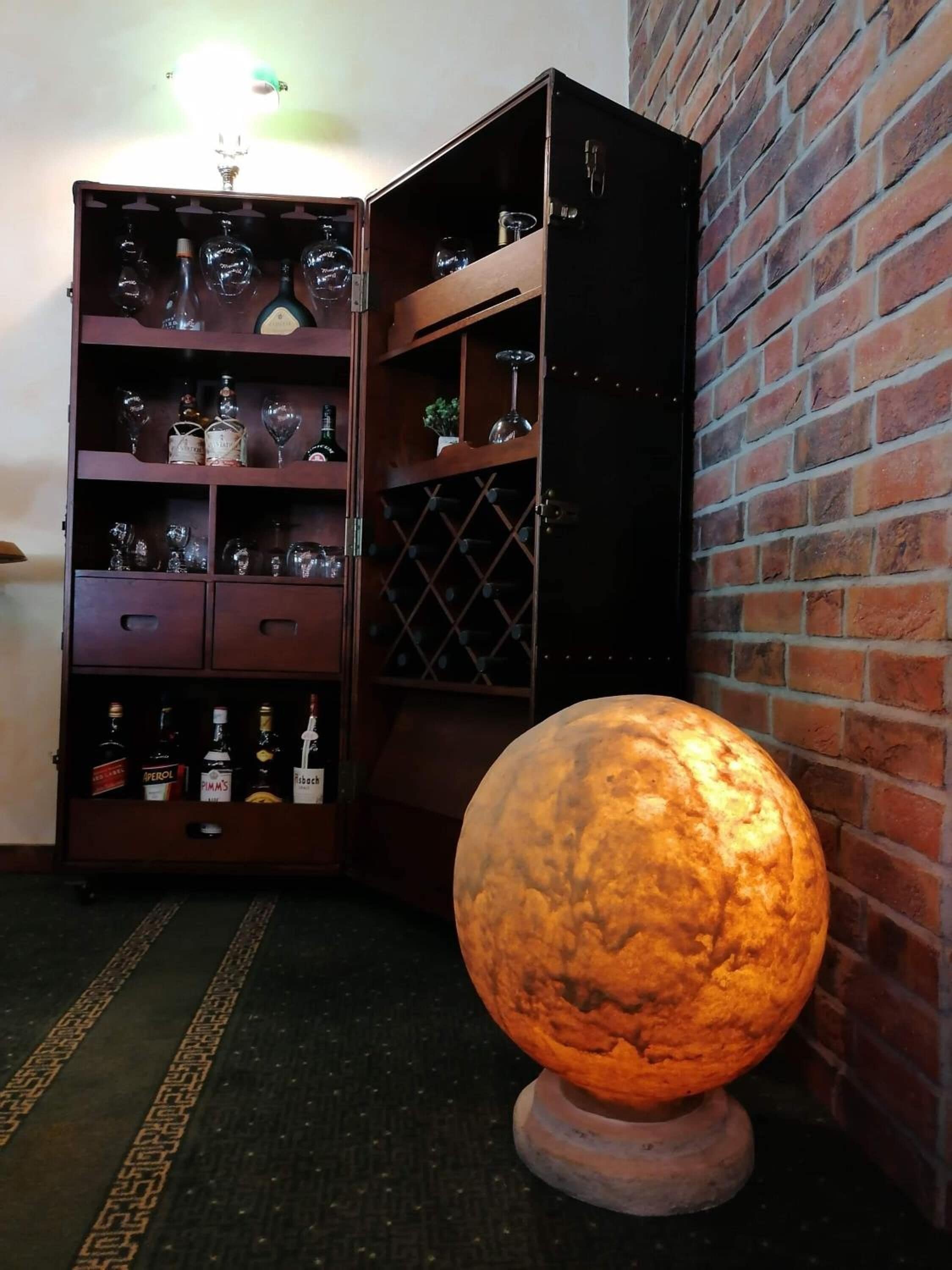 A corner with a brick wall, a wooden wine rack with bottles and glasses, and a large illuminated globe.