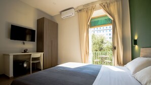 A hotel room with a bed, a desk, a chair, a TV, an air conditioner, and a balcony with a view of buildings and trees. - Interno 10 Cavour (Naples)