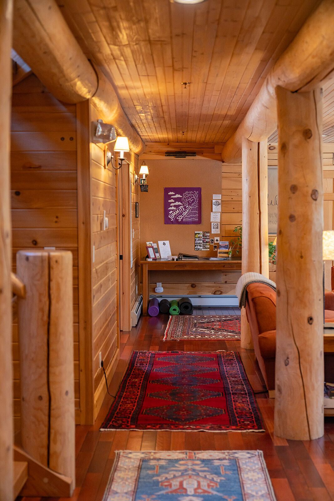 Een houten gang met een vloerkleed in rood en blauw patroon, houten wanden en een bankje.