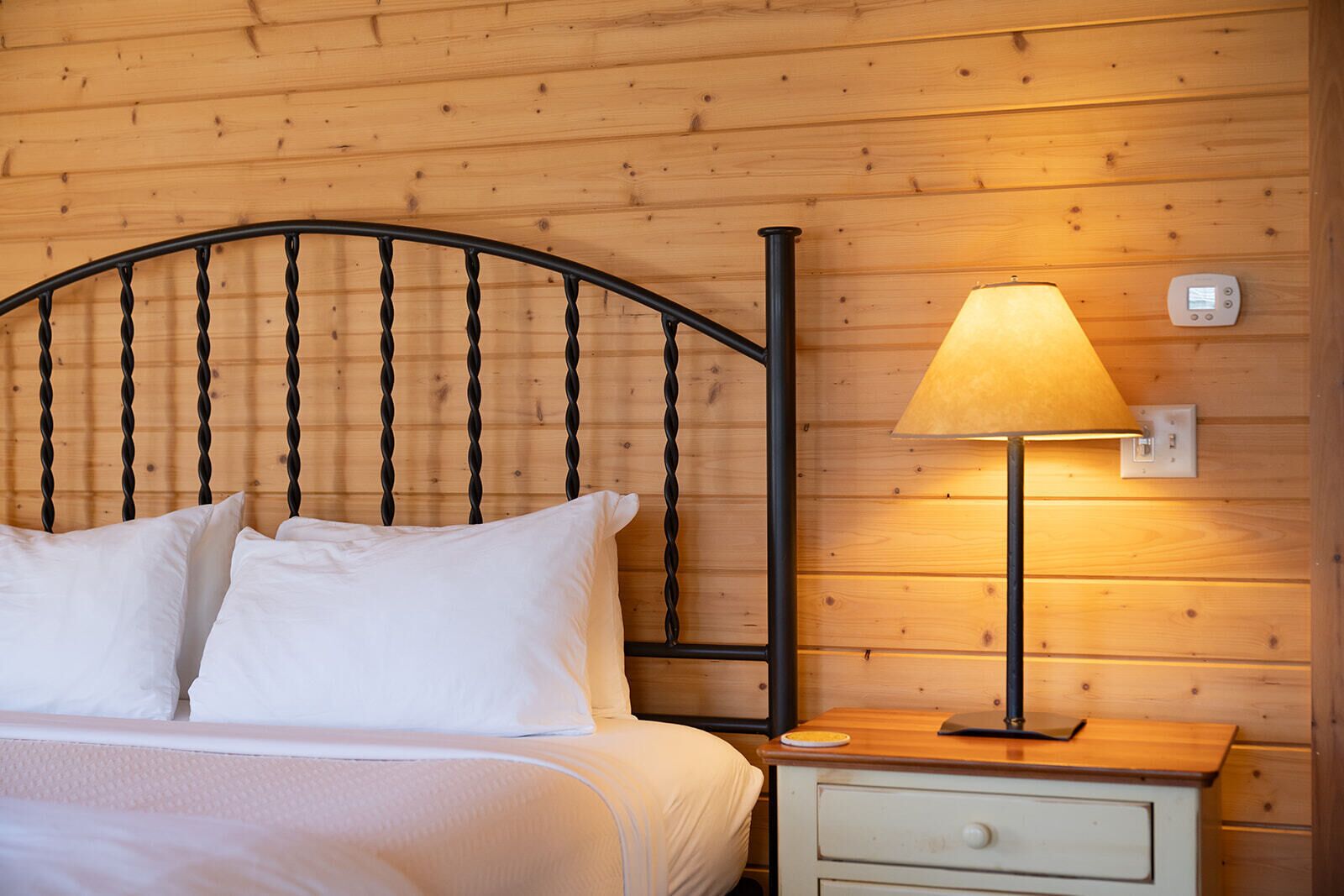 A bed with white linens, a black metal headboard, a bedside lamp, and a small white nightstand with a drawer.