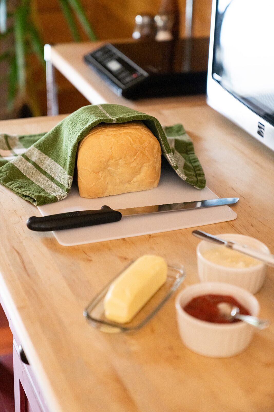 A loaf of bread on a cutting board with a knife, a butter dish, and a bowl of jam.