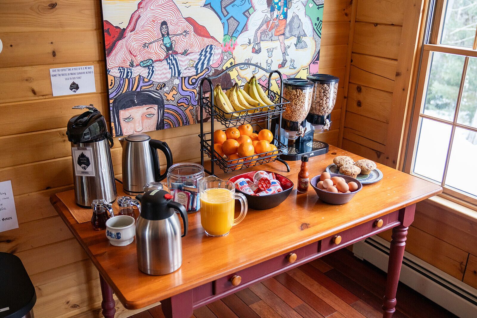 A wooden table with breakfast items including coffee, eggs, bananas, oranges, and muffins.