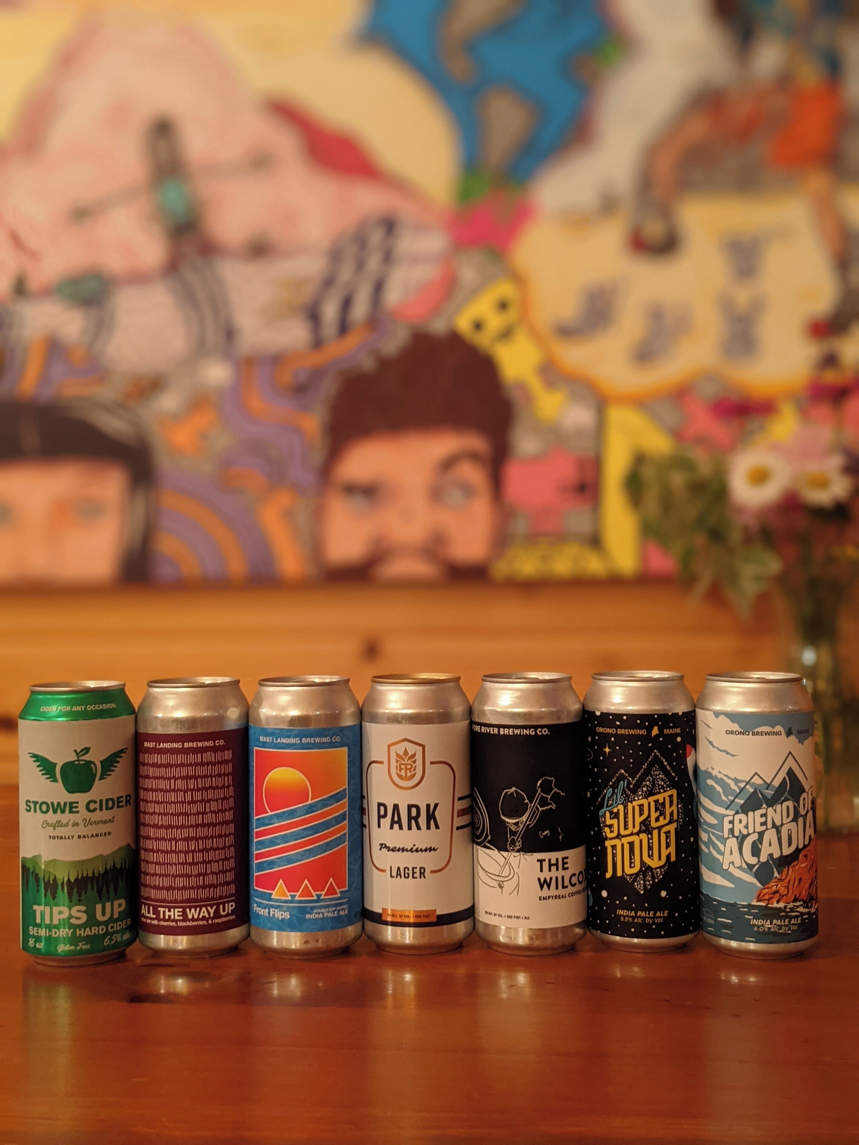 A row of beer cans on a wooden surface with a colorful mural in the background.