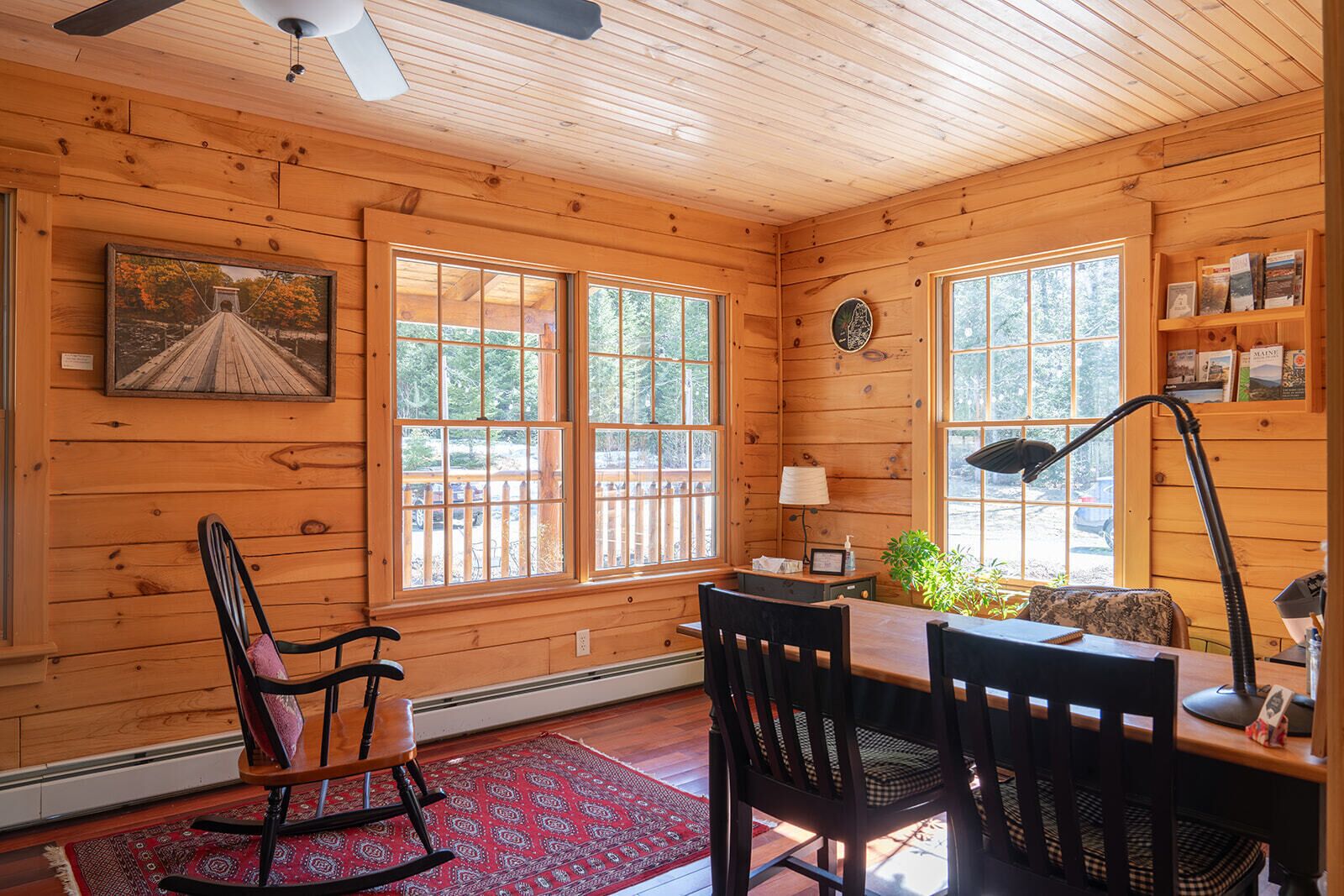 L’intérieur chaleureux d’un chalet avec des murs en bois, de grandes fenêtres, un fauteuil à bascule, un bureau avec une lampe et une bibliothèque.