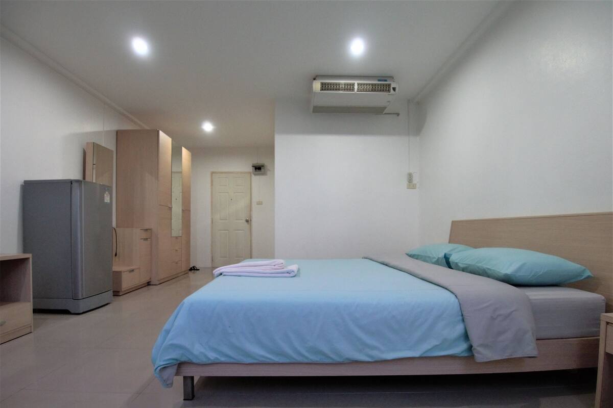 A bedroom with a bed, a nightstand, a wardrobe, a refrigerator, and an air conditioning unit.
