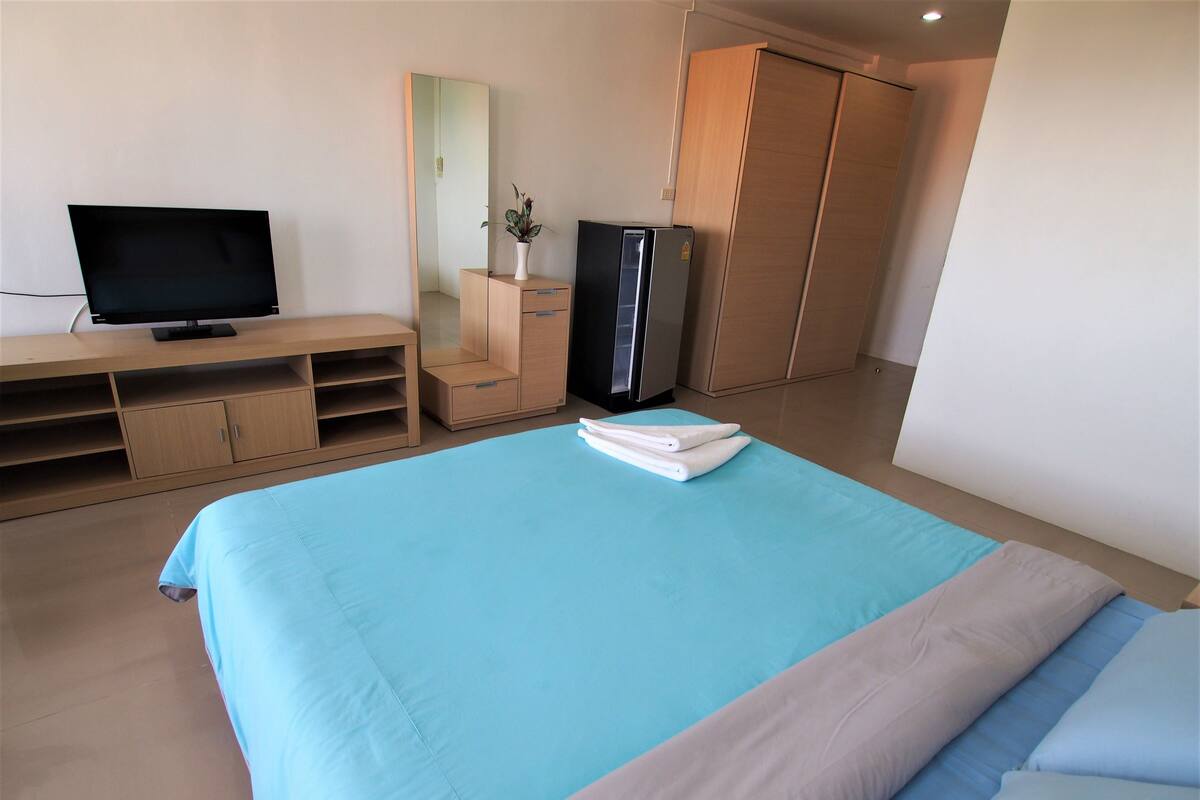 A hotel room with a bed, a TV, a wardrobe, and a desk with a mirror and a plant.