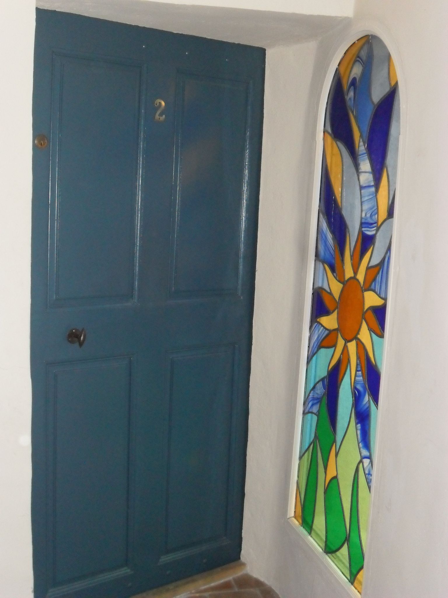 Une porte bleue avec une poignée de porte en laiton et un vitrail représentant un motif solaire.