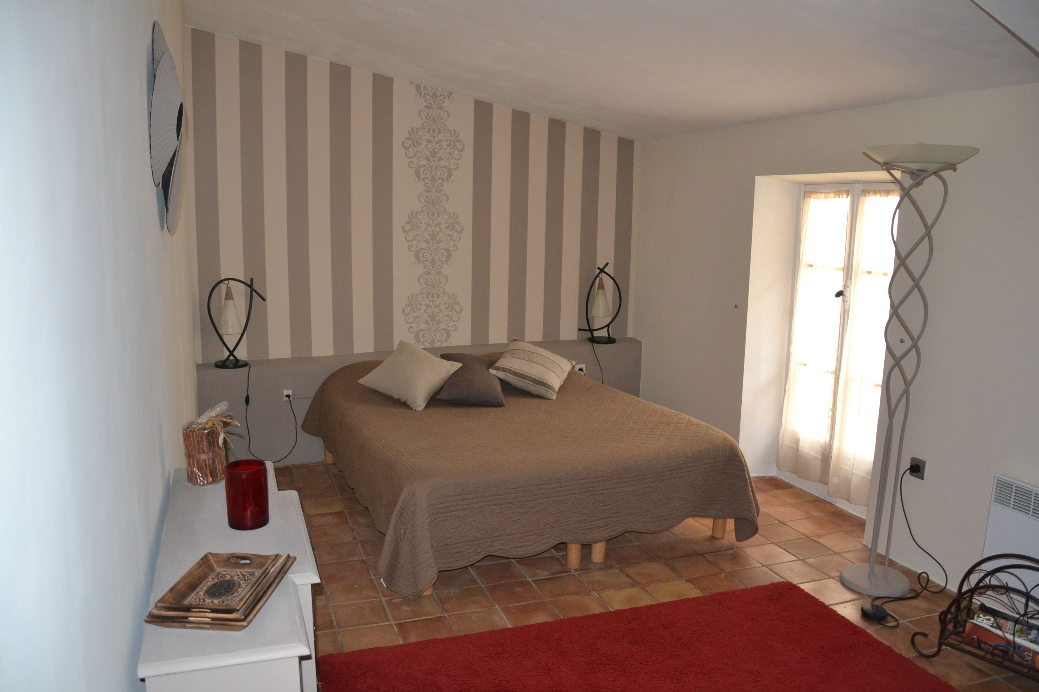 A bedroom with a bed, a bedside table, a red rug, a floor lamp, and a window with curtains.