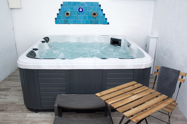 A hot tub with a wooden bench and a small stool, surrounded by a tiled wall and a white wall.