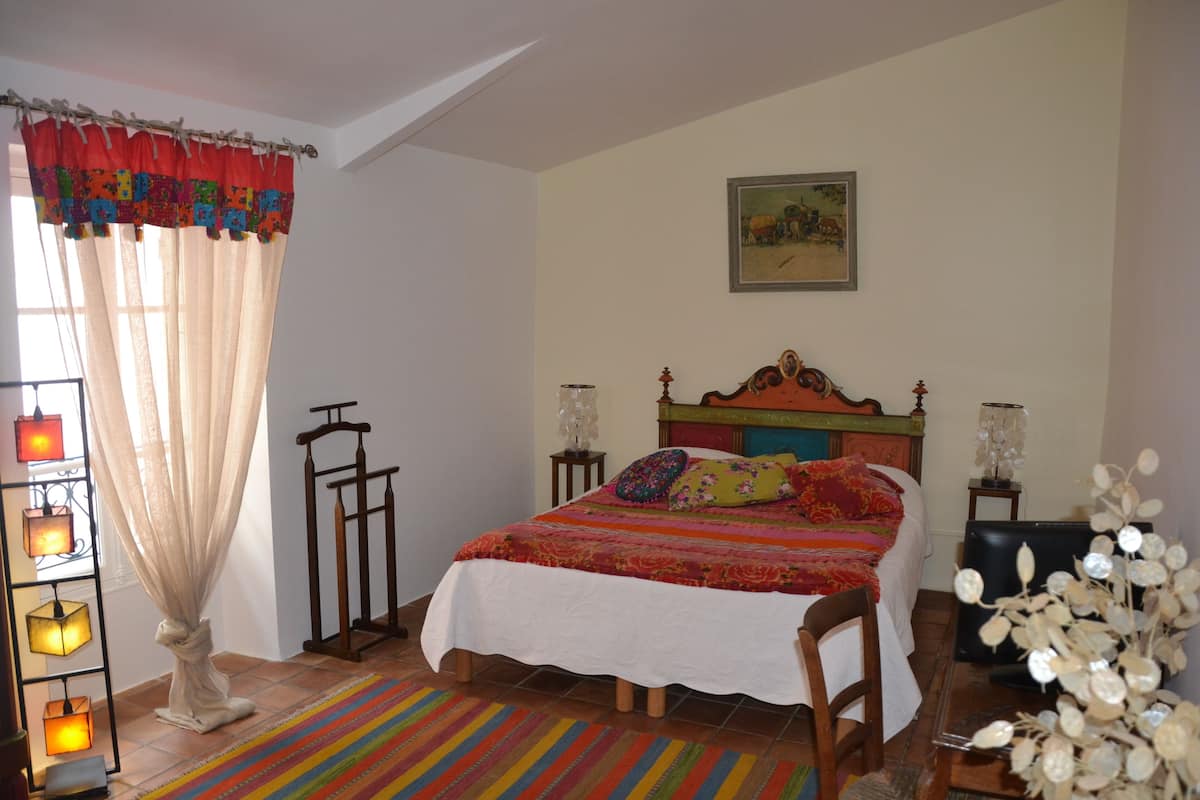 Ein Schlafzimmer mit einem Bett, Nachttischlampen, einem Nachttisch, einem Sessel, einem Fenster mit Vorhängen und einem an der Wand hängenden Bild.