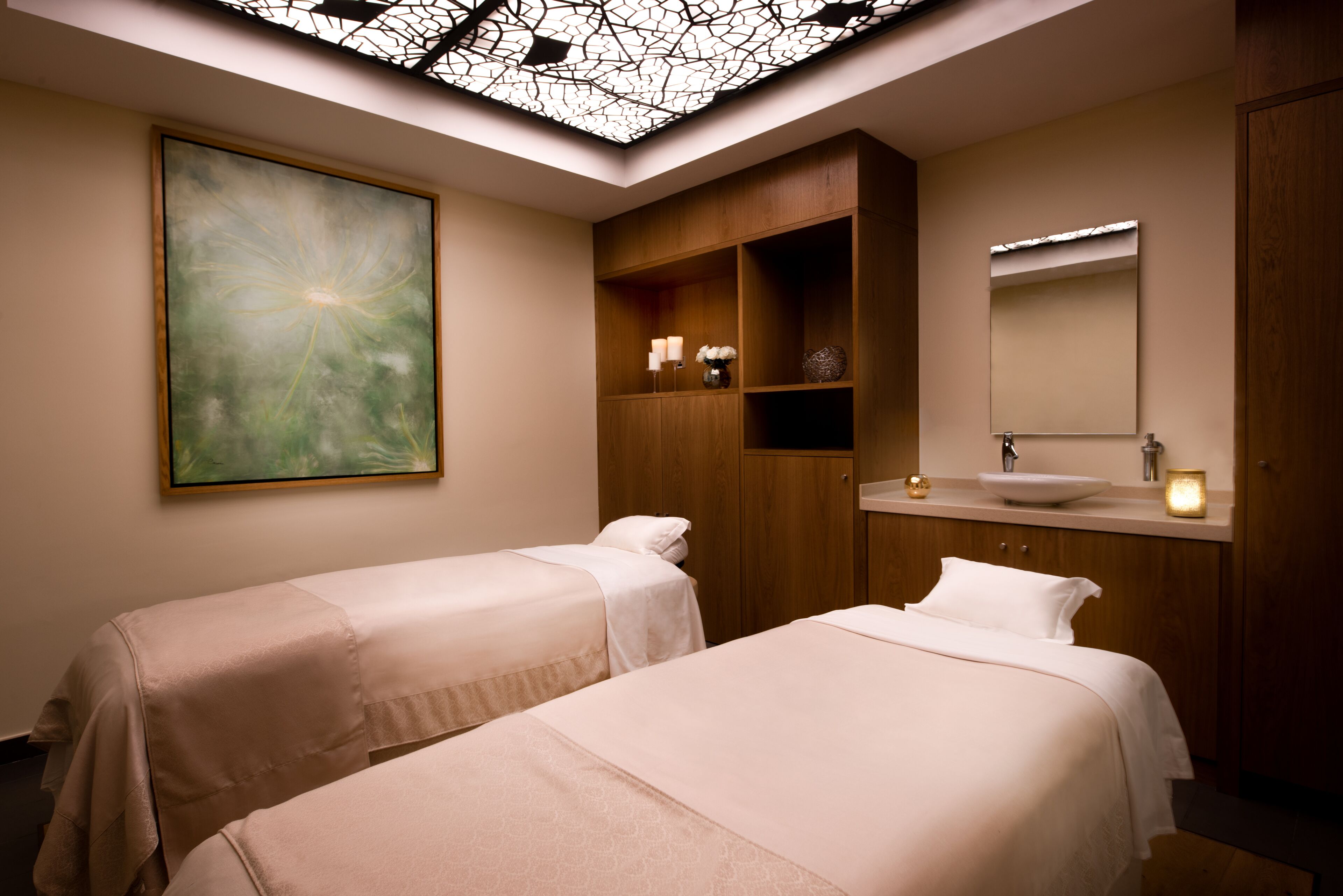 A hotel room with two beds, a large abstract painting, a wooden cabinet, a vanity with a mirror, and a skylight ceiling.