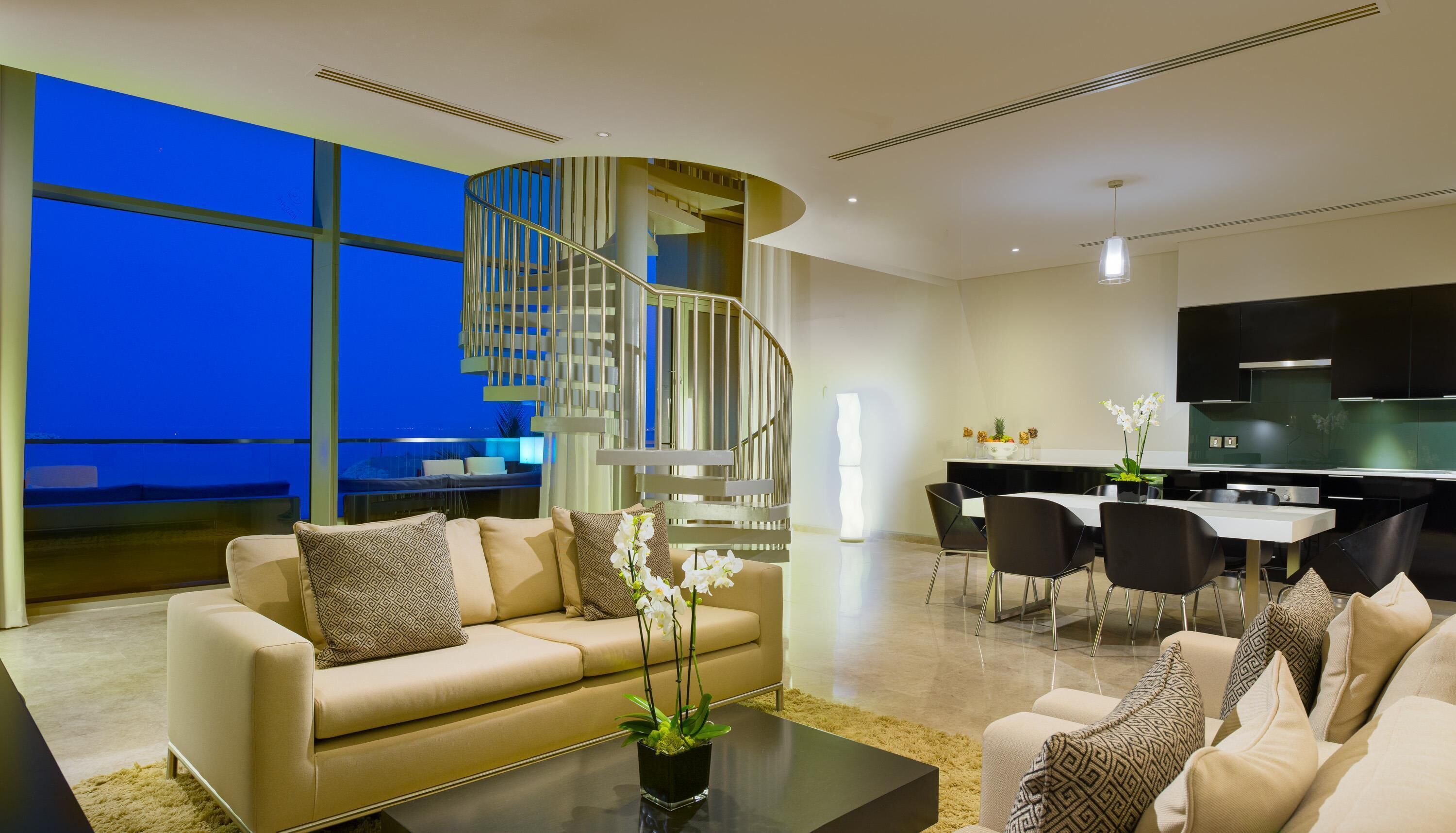 A modern living room with a curved staircase, a dining area, and a kitchen with a large TV.