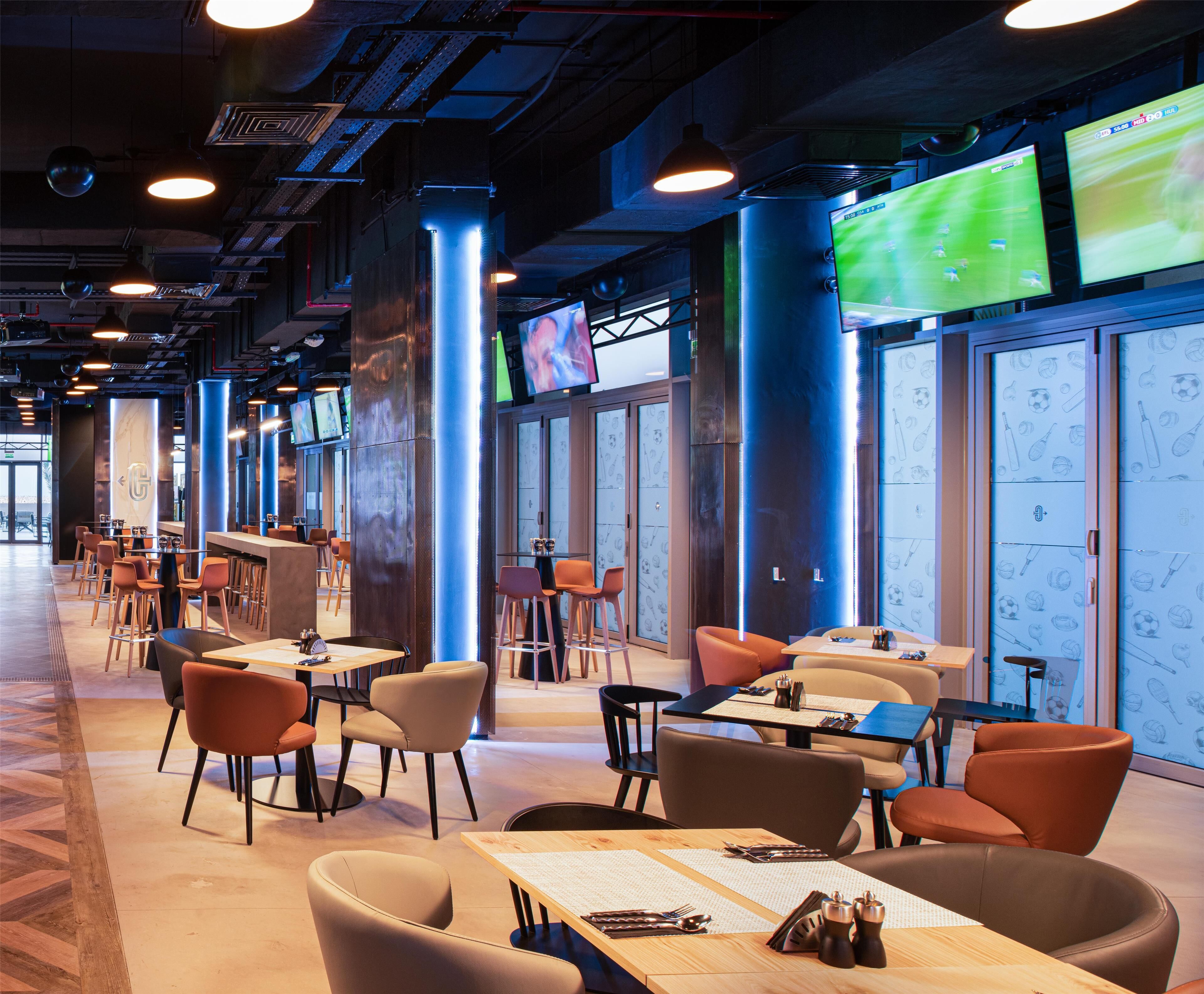 A modern indoor space with multiple seating areas, including tables and chairs, and large screens displaying sports events.