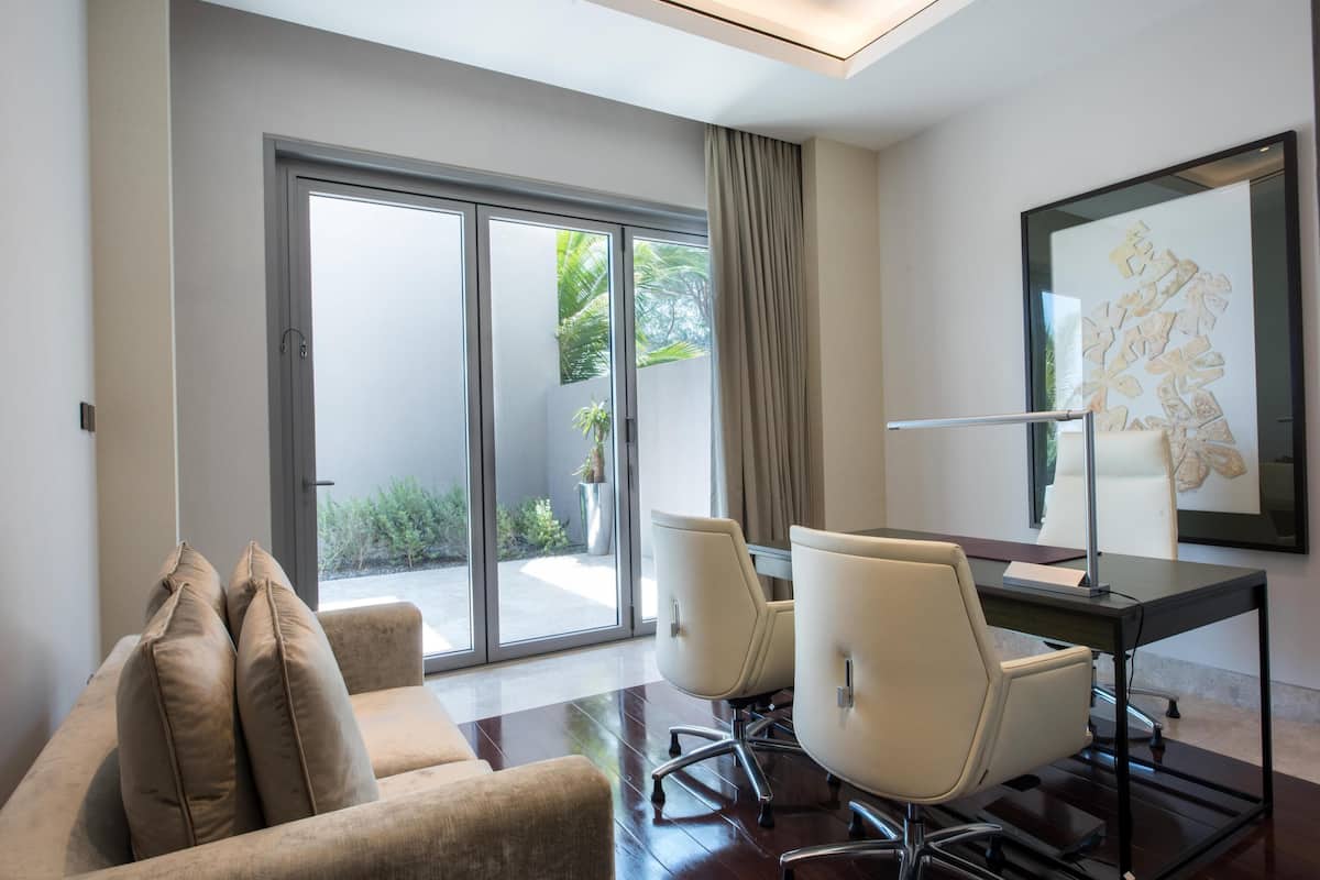 A modern room with a sofa, a desk with two chairs, a large window with a view of greenery, and a framed artwork on the wall.