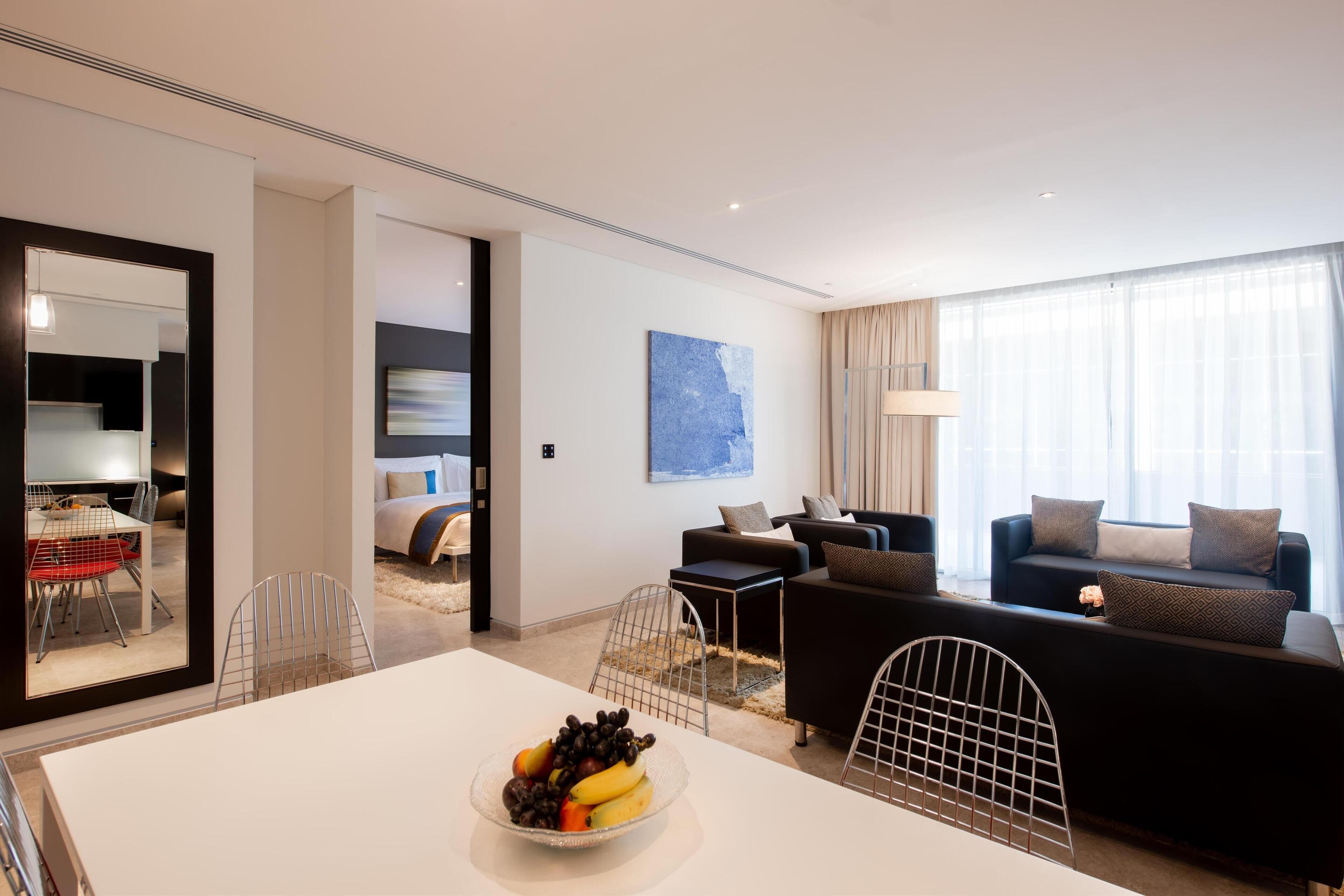 A modern hotel room with a dining area, a living space with a sofa and a coffee table, and a large window with curtains.