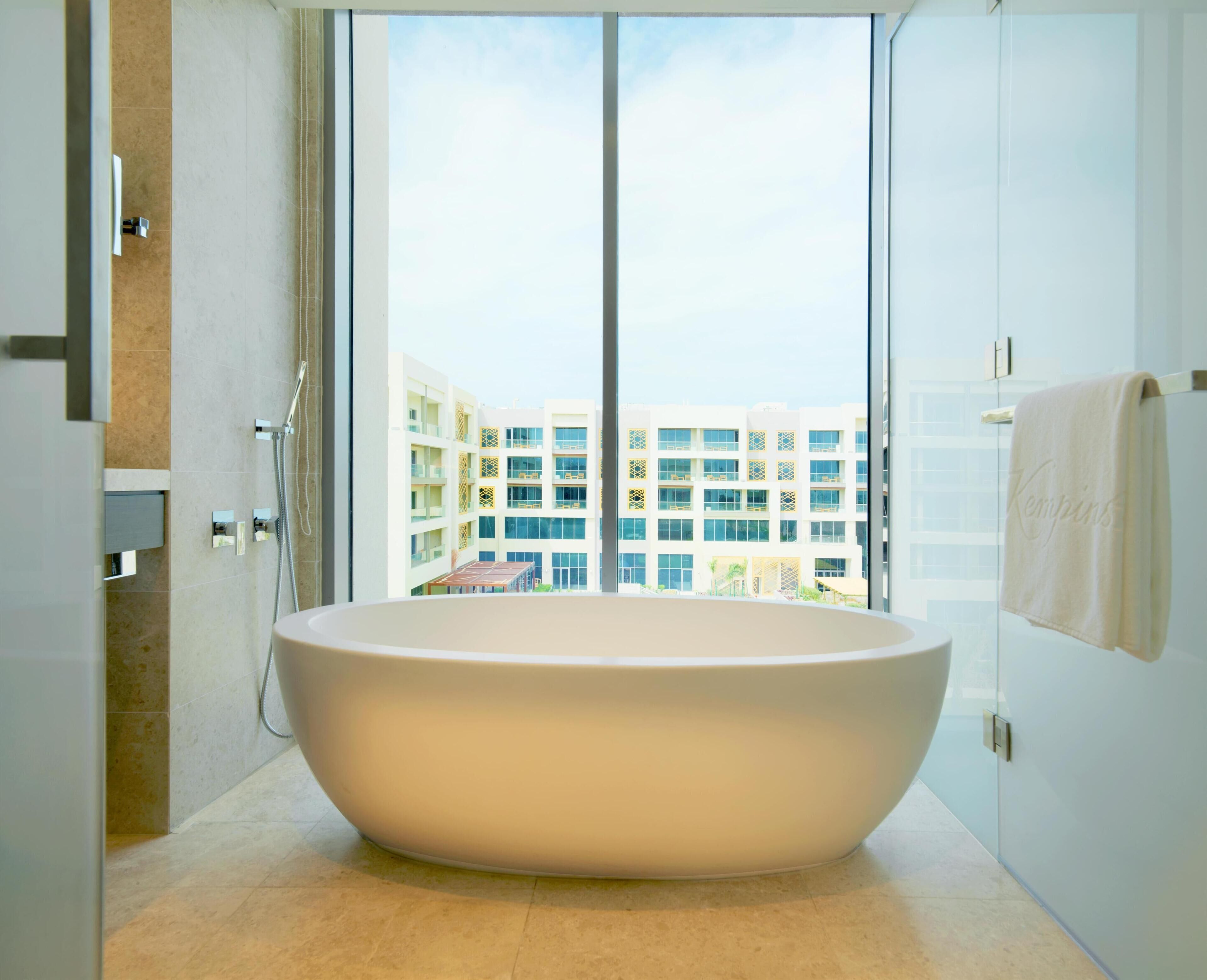 A modern bathroom with a large freestanding bathtub, a glass shower enclosure, and a view of a multi-story building through a large window.