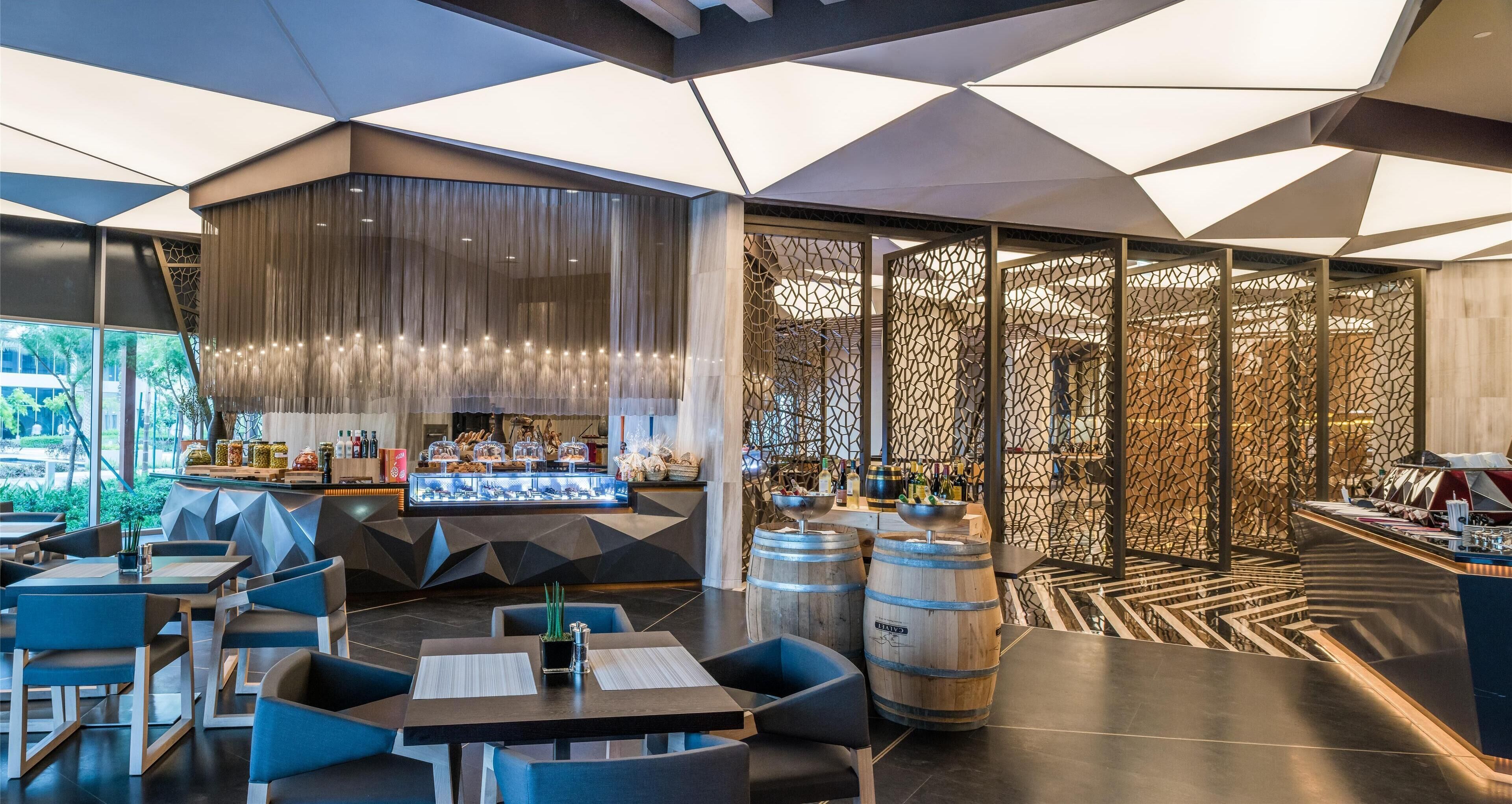 Un restaurant moderne doté d’un plafond au design géométrique, d’un bar où sont exposées des bouteilles et d’un espace détente avec des tables et des chaises.