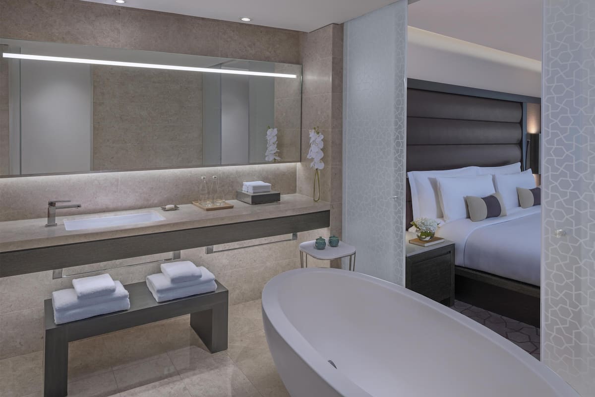 A modern bathroom with a large mirror, a bathtub, a vanity with a sink, and a bedside table with a lamp.