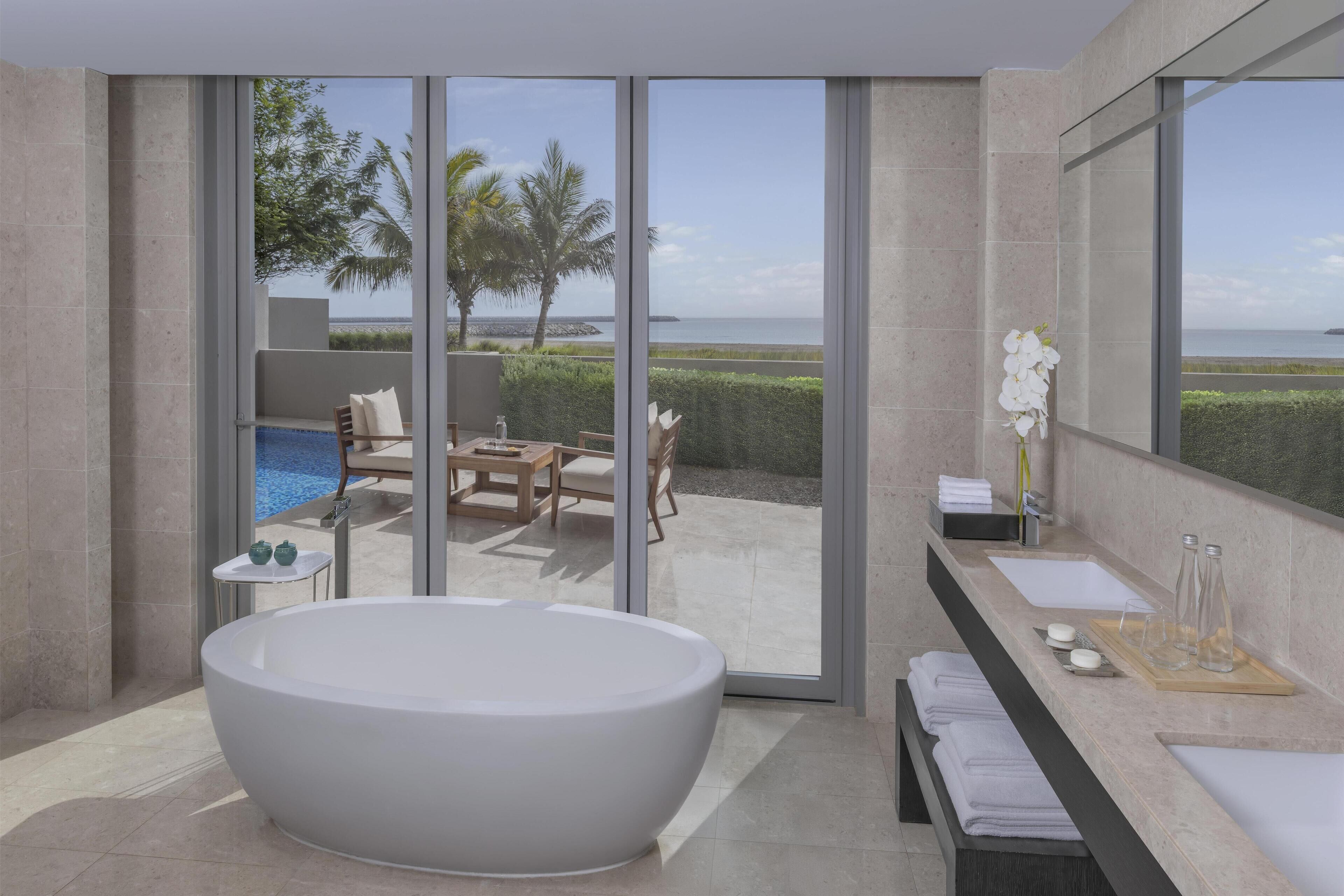 Une salle de bain moderne avec une grande baignoire, un lavabo et un meuble-lavabo. De grandes portes vitrées offrent une vue sur l’océan et les palmiers.