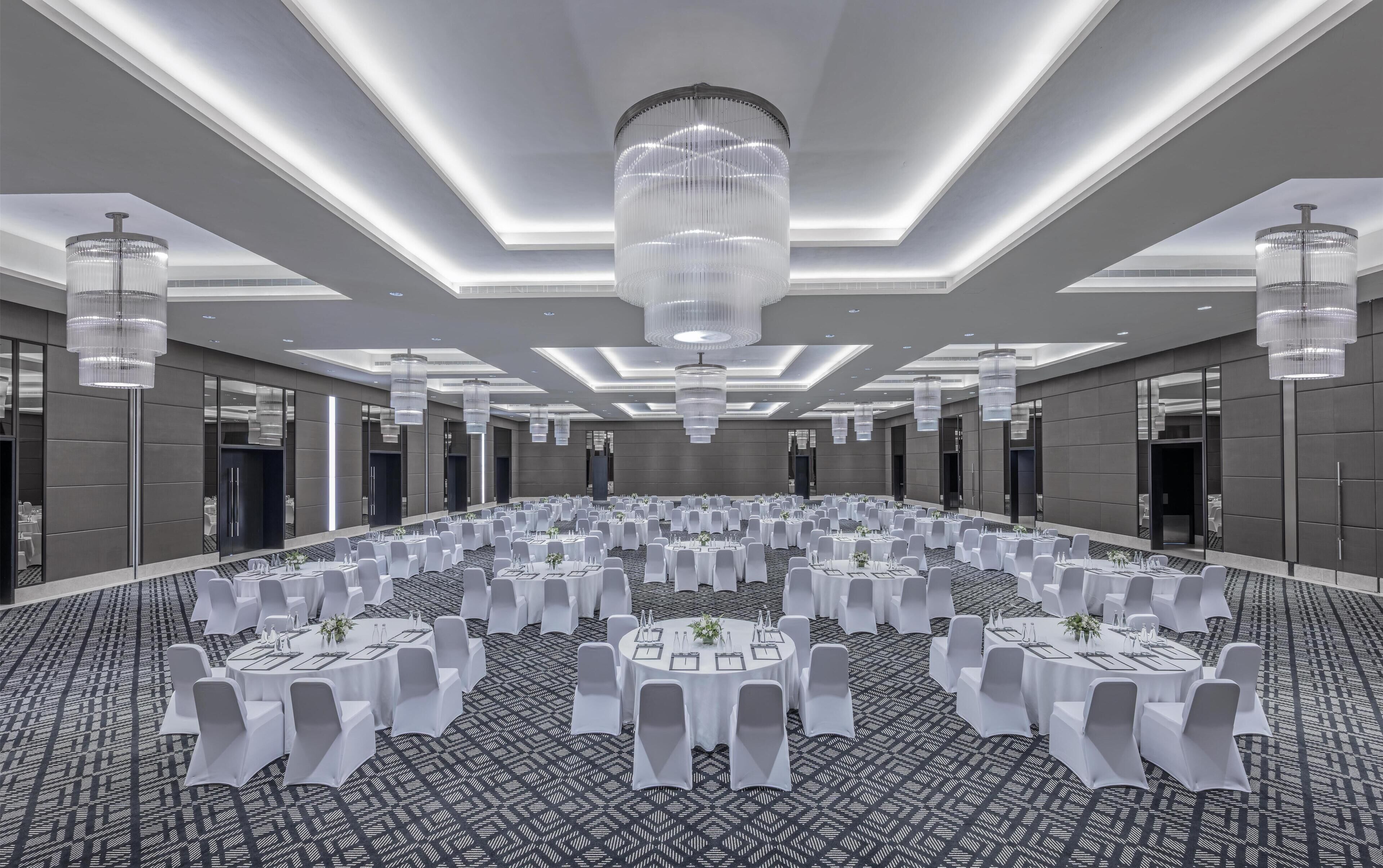 Un amplio salón de conferencias con mesas redondas, sillas blancas y una alfombra estampada.